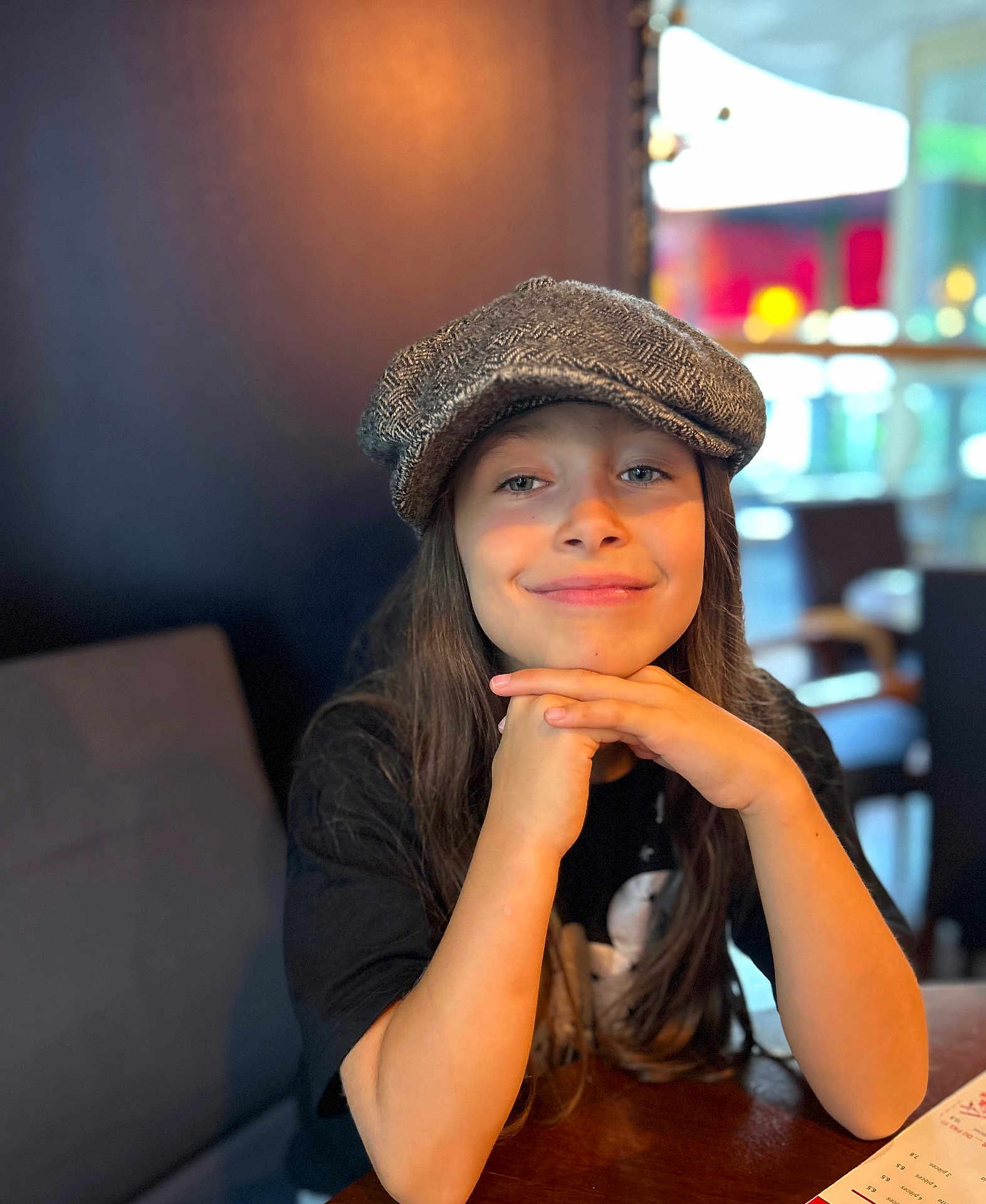 Tessa Lou a rejoint le concours — aidez-le/la à gagner de superbes lots ! child, smile, hat, cap, long_hair, face, portrait, indoor, table, wood, clothing, person, seated, casual, young, happy, restaurant, blurred_background, fashion, hands