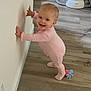 baby, child, pink_onesie, smiling, standing, wooden_floor, indoor, wall, baby_swing, toy, footwear, cute, happy, person, infant, home, flooring, looking_at_camera, small_toy, balance