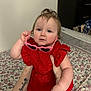 baby, child, red_dress, heart_shaped_sunglasses, tattoo, indoor, bed, floral_bedding, person, cute, blue_eyes, curly_hair, hand, arm, holding, portrait, infant, playful, fashion, accessory