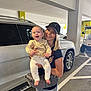 adult, baby, cap, car, casual, child, clothing, expression, footwear, happy, holding, jeans, light, parking_garage, person, smile, urban, vehicle, white_suv, woman