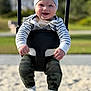 Mark is registered to the contest to win money with this photo: baby, child, swing, smiling, hat, striped_clothing, camouflage_pants, socks, playground, sand, outdoor, daylight, happy, toddler, person, seat, fun, nature, blurred_background, casual_clothing