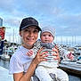baby, baby_carrier, boats, cap, casual_clothing, child, cloudy, daylight, dock, happy, hat, holding, marina, outdoor, person, sky, smile, striped_clothing, water, woman