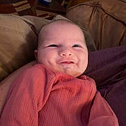 Bailey is registered to the contest to win money with this photo: baby, smile, infant, pink_clothing, couch, indoor, face, person, child, cheeks, cozy, furniture, relaxing, home, portrait, cute, sitting, happy, young, expression
