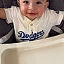 baby, child, high_chair, dodgers, smiling, face, food_smudge, clothing, person, indoor, seat, happy, cute, infant, portrait, table, messy, baseball, sportswear, young
