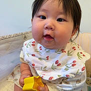 Monako is registered to the contest to win money with this photo: baby, child, infant, face, hand, toy, yellow, bib, hair_clip, indoor, cute, chubby_cheeks, clothing, sitting, play, curious, skin, expression, closeup, portrait