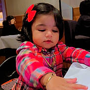 Kaylee joined the competition — help win amazing prizes! toddler, child, girl, red_bow, plaid_dress, jewelry, table, restaurant, indoor, curious, black_hair, high_chair, person, portrait, cute, baby, dining, background_people, wall_decor, colorful_clothing