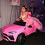 child, toddler, pink_dress, toy_car, birthday_party, balloons, celebration, happy, smiling, indoor, event, decorations, fairy_theme, dress_up, vehicle, small_car, girl, party, joyful, fun