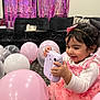 child, toddler, birthday_party, balloons, pink_dress, headband, camera, indoor, celebration, happy, smiling, decorations, banner, pink, purple, toy, curly_hair, carpet, couch, party