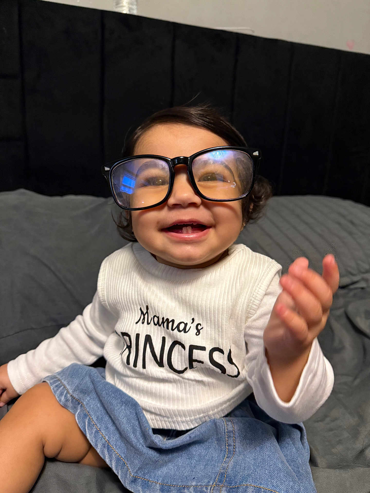 Sanaya is registered to the contest to win money with this photo: apparel, baby, bed, bedding, child, clothing, cute, denim_skirt, glasses, happy, headboard, indoor, infant, joyful, person, portrait, sitting, smiling, white_shirt, young_child