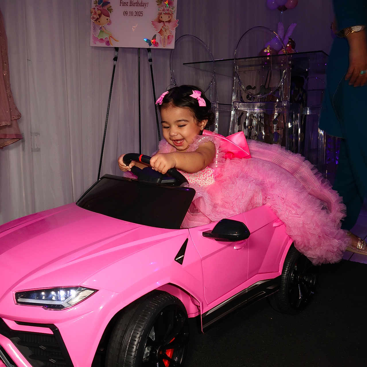 Sanaya is registered to the contest to win money with this photo: balloons, birthday_party, celebration, child, clothing, decorations, event, face, floor, fun, happy, indoor, joy, person, pink_dress, ribbons, smiling, steering_wheel, toddler, toy_car