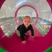 Olivier is registered to the contest to win money with this photo: toddler, child, playground, play_tunnel, crawling, indoor_play_area, pink, green, smiling, curious, plastic, circular, netting, fun, adventure, young_child, clothing, black_shirt, gray_pants, hands