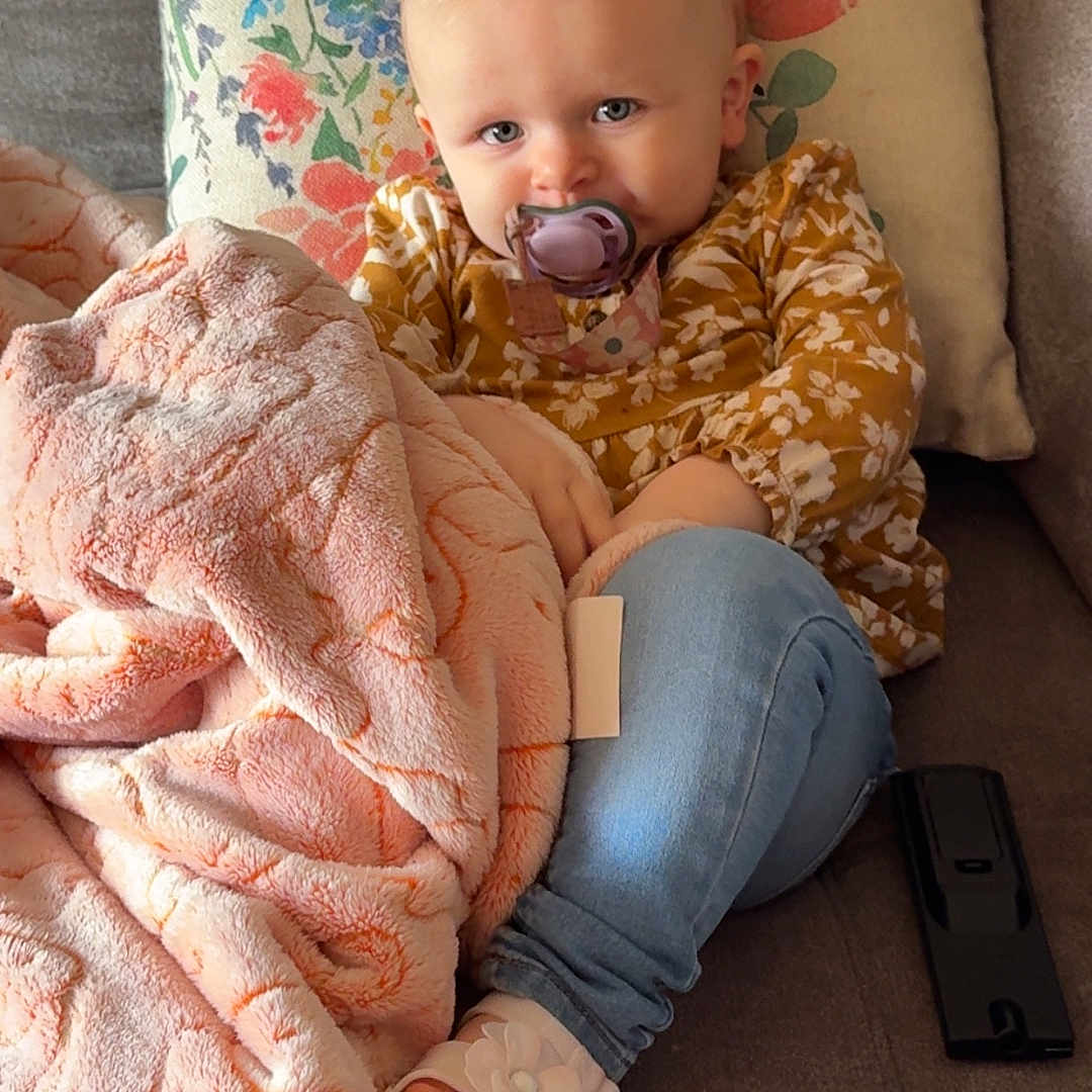 Enora joined the competition — help win amazing prizes! toddler, child, pacifier, blanket, couch, pillow, floral_pattern, yellow_top, blue_jeans, white_sandals, flower_decoration, indoor, cozy, cute, sitting, curious, soft_texture, home, relaxing, person