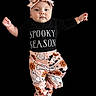 baby, infant, child, portrait, spooky_season, headband, bow, black_clothing, patterned_pants, floral_pattern, ghost_pattern, arms_outstretched, floating_pose, bare_feet, facial_expression, face, indoor_studio, black_background, fashion, photography