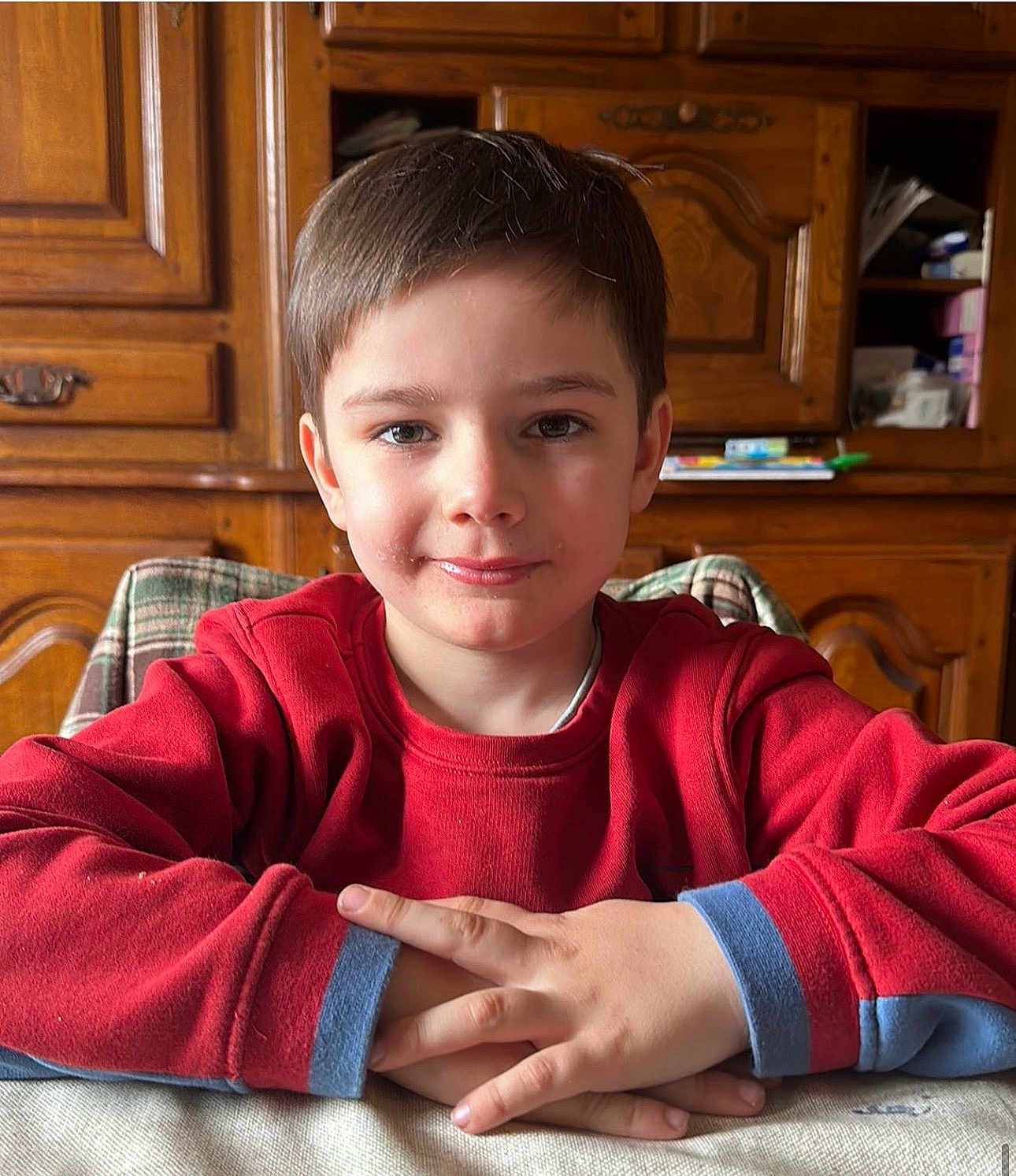Adrien a rejoint le concours — aidez-le/la à gagner de superbes lots ! child, boy, red_sweatshirt, hands, smile, face, table, furniture, wooden_cabinet, indoor, portrait, casual, cozy, young, person, home, sitting, friendly, natural_light, relaxed