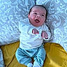 baby, infant, smiling, happy, clothing, pillow, blanket, bed, lying_down, person, cute, blue_pants, white_shirt, socks, indoor, patterned_pillow, yellow_blanket, small_hands, face, joyful