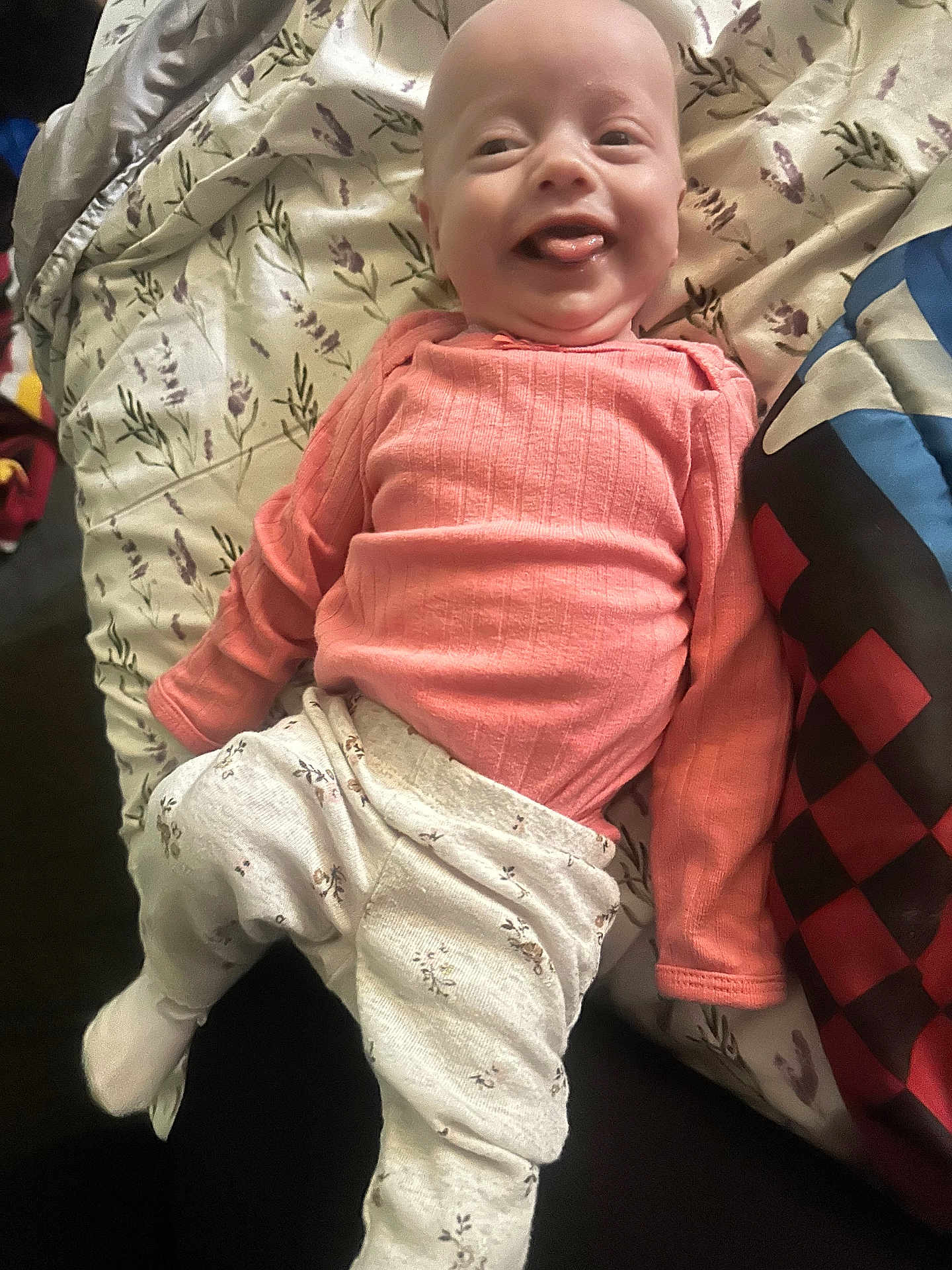 Paisleigh joined the competition — help win amazing prizes! baby, infant, smiling, tongue_out, pink_sweater, floral_pants, blanket, patterned_blanket, lying_down, happy, cute, child, indoors, cozy, fabric, soft_lighting, portrait, young_child, person, cheerful