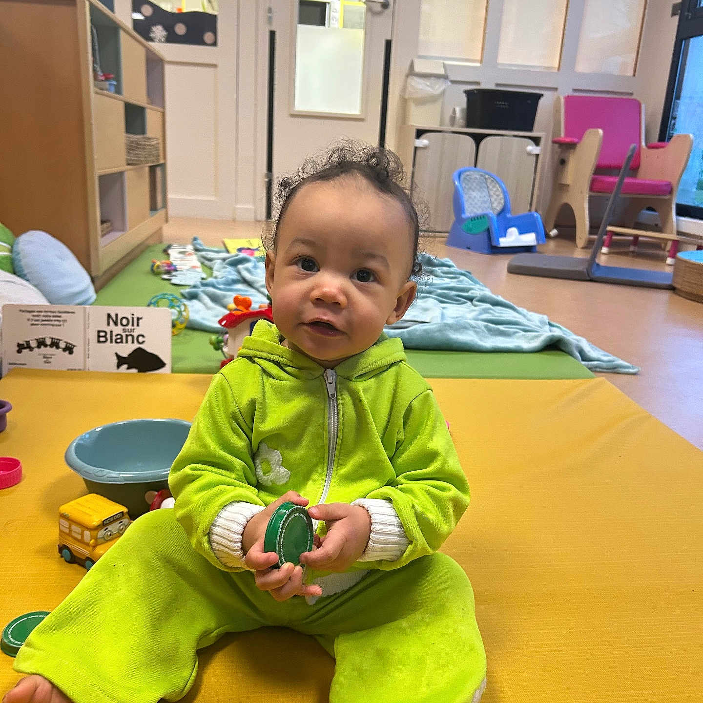 Jaelya participe au concours pour gagner de l'argent avec cette photo : baby, blanket, bowl, chair, child, curly_hair, door, floor, furniture, green_clothing, indoor, person, plastic_ring, play, playroom, smiling, toddler, toy, window, yellow_mat