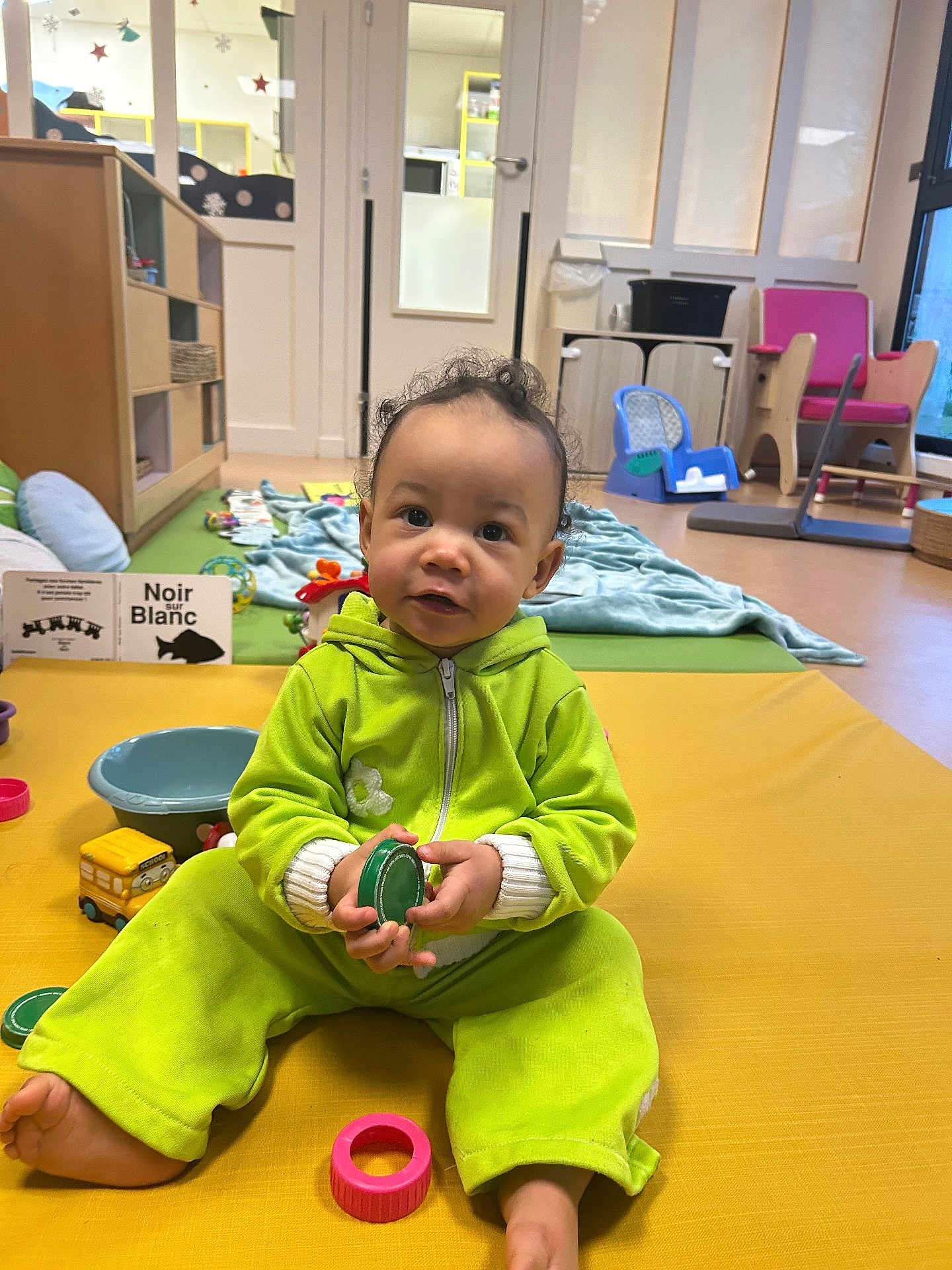 Jaelya participe au concours pour gagner de l'argent avec cette photo : baby, child, toddler, playroom, toy, plastic_ring, green_clothing, yellow_mat, floor, indoor, furniture, blanket, window, door, chair, bowl, smiling, curly_hair, person, play