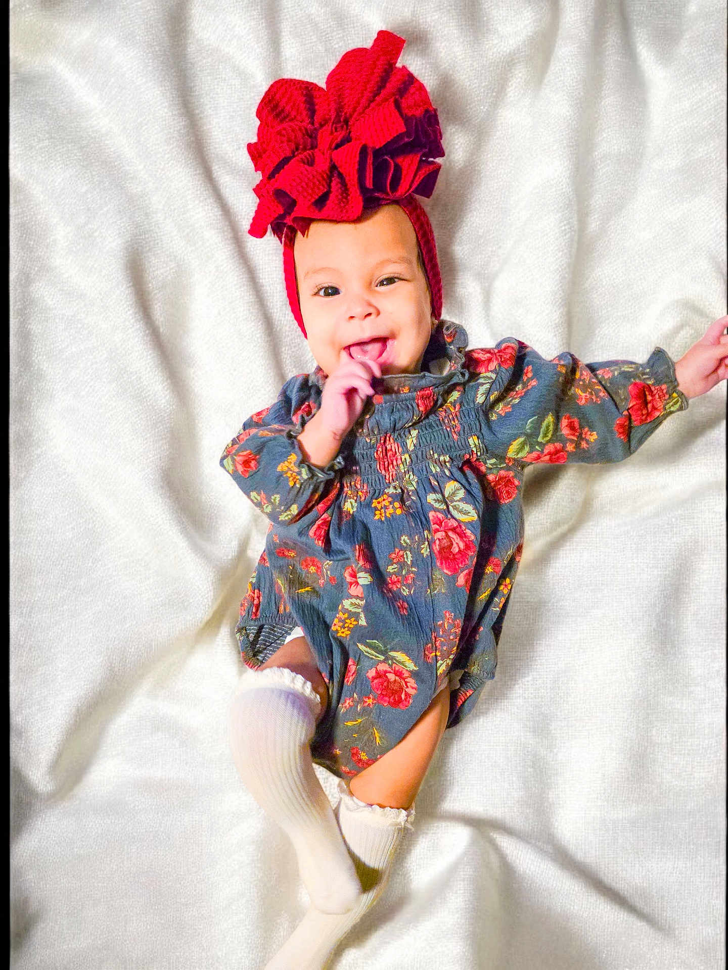 Evangeline joined the competition — help win amazing prizes! baby, child, smiling, headband, red_bow, floral_dress, white_socks, blanket, lying_down, happy, cute, infant, one_hand_up, playful, indoors, portrait, soft_texture, colorful_clothing, small_child, person