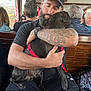 man, dog, hug, beard, tattoo, cap, sunglasses, bench, passengers, vehicle, window, red_harness, black_dog, casual_clothing, shorts, watch, indoor, people, tattooed_arm, affection