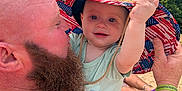 Killian is registered to the contest to win money with this photo: baby, man, beard, tattoo, hat, straw_hat, outdoor, trees, sand, sunny, child, person, smile, arm, green_clothing, bracelet, nature, daylight, closeup, holding