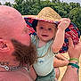 baby, man, beard, tattoo, hat, straw_hat, outdoor, trees, sand, sunny, child, person, smile, arm, green_clothing, bracelet, nature, daylight, closeup, holding