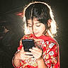 child, girl, phone, red_clothing, floral_pattern, backlight, dark_background, portrait, technology, engaged, hair, hands, traditional_clothing, indoors, casual, young, ethnic_wear, patterned, concentration, light