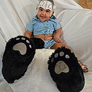 Zoe is registered to the contest to win money with this photo: baby, child, smiling, helmet, footwear, oversized_slippers, bear_paw_slippers, plush, white_background, blue_clothing, sitting, happy, cute, indoor, portrait, person, furry, soft_texture, headgear, footwear_closeup