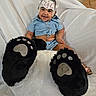 baby, child, smiling, helmet, footwear, oversized_slippers, bear_paw_slippers, plush, white_background, blue_clothing, sitting, happy, cute, indoor, portrait, person, furry, soft_texture, headgear, footwear_closeup