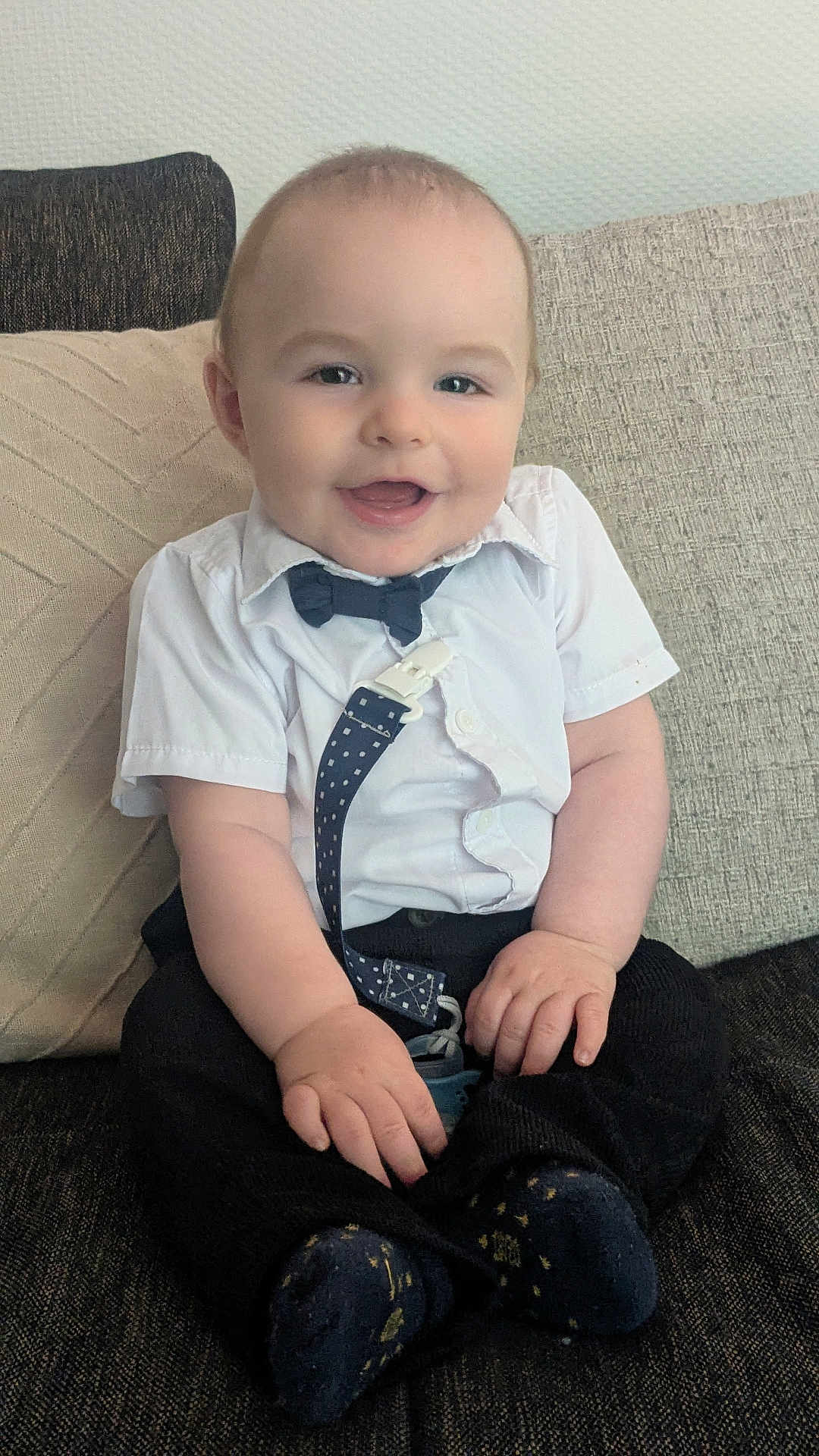 Ophélien participe au concours pour gagner de l'argent avec cette photo : baby, child, smiling, sitting, shirt, bow_tie, suspenders, couch, cushion, happy, indoors, face, person, cute, infant, clothing, footwear, sock, home, portrait