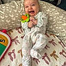 baby, infant, smiling, toy, onesie, pillow, heart_pattern, blanket, cap, indoor, cute, happy, child, face, person, soft_toys, playful, cozy, lying_down, comfortable