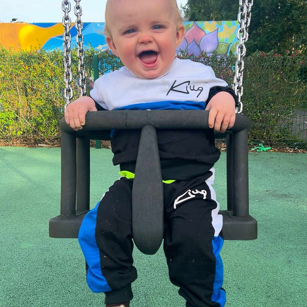 Jayden joined the competition — help win amazing prizes! bear, chains, child, clothing, colorful, daylight, fence, fun, greenery, happy, mural, outdoor, park, person, playground, sky, smiling, socks, swing, toddler