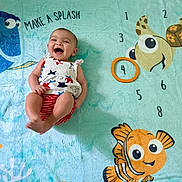 Selena is registered to the contest to win money with this photo: baby, blanket, cartoon_fish, child, clothing, colorful, cute, fun, happy, infant, laughing, lying_down, marine_life, patterned, playful, red_shorts, smiling, star_pattern, turtle, underwater_theme