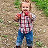 child, toddler, smiling, outdoors, apples, fruit, orchard, dirt_path, grass, nature, portrait, standing, plaid_shirt, jeans, sneakers, ponytail, happy, red_apples, facial_expression, daylight