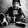 child, toddler, boy, baseball_cap, jacket, jeans, sneakers, toy_car, sippy_cup, portrait, black_and_white, studio, wooden_floor, seated, serious_expression, direct_gaze, casual_style, close_up, beverage_container, cool_tshirt