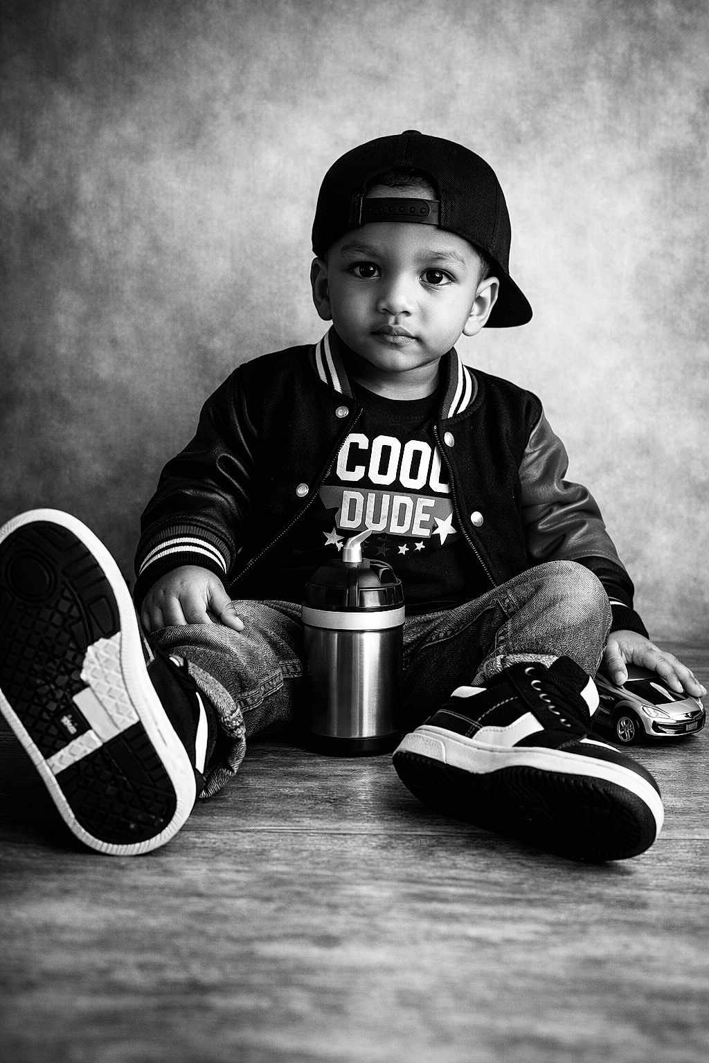 Lucienzo a rejoint le concours — aidez-le/la à gagner de superbes lots ! child, toddler, boy, baseball_cap, jacket, jeans, sneakers, toy_car, sippy_cup, portrait, black_and_white, studio, wooden_floor, seated, serious_expression, direct_gaze, casual_style, close_up, beverage_container, cool_tshirt