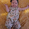 Jadzia is registered to the contest to win money with this photo: baby, child, infant, smiling, portrait, lying_down, floral_blanket, yellow, dress, headband, bow, indoors, closeup, face, arms, feet, patterned_clothing, happy, bed, photograph