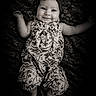 baby, child, infant, smiling, portrait, black_and_white, monochrome, lying_down, floral_dress, headband, bow, blanket, patterned_background, outstretched_arms, bare_feet, indoor, studio_photography, happy, face, vintage_style
