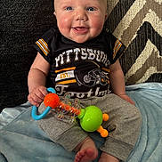 Jaxon is registered to the contest to win money with this photo: baby, smiling, blue_eyes, rosy_cheeks, pittsburgh_football_shirt, gray_pants, rattle, toy, sitting, blanket, blue_blanket, patterned_rug, indoor, child, cute, happy, infant, person, footwearless, closeup
