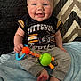 baby, smiling, blue_eyes, rosy_cheeks, pittsburgh_football_shirt, gray_pants, rattle, toy, sitting, blanket, blue_blanket, patterned_rug, indoor, child, cute, happy, infant, person, footwearless, closeup