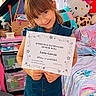 Luna is registered to the contest to win money with this photo: child, girl, smiling, portrait, honor_roll, certificate, award, bedroom, hello_kitty, stuffed_animal, school, student, blue_dress, standing, indoors, window, curtains, colorful_bedding, books, happy