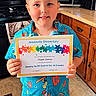 child, boy, portrait, smiling, certificate, award, achievement, elementary_school, standing, kitchen, wooden_cabinets, stove, microwave, countertop, colorful_shirt, floral_shirt, shorts, indoors, proud, family_photo