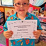 child, boy, glasses, smiling, portrait, certificate, award, honor_roll, school, student, standing, indoors, colorful_shirt, tropical_pattern, toy_room, window, furniture, carpet, proud, achievement