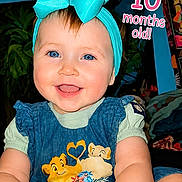 Amara joined the competition — help win amazing prizes! baby, infant, child, portrait, smiling, tongue_out, blue_eyes, bow, headband, turquoise, dress, disney, lion_king, sitting, indoor, plants, shelf, bottles, milestone, text_overlay