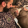 man, dog, bed, tattoo, beard, close_up, indoor, resting, patterned_clothing, pillow, pet, companion, relaxed, casual, portrait, human, animal, friendship, comfort, affection