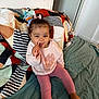 Arya participe au concours pour gagner de l'argent avec cette photo : toddler, child, smiling, sitting, blanket, patterned_blanket, indoors, toy, hair_bow, pink_clothing, leggings, sweatshirt, barefoot, floor, door, wall, playful, happy, face, person