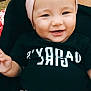 apparel, baby, black_shirt, child, cute, face, fashion, girl, happy, headband, human, infant, outdoor, person, pink, portrait, sitting, skin, smiling, text_on_clothing