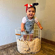 Lucy joined the competition — help win amazing prizes! baby, child, smiling, overalls, headband, basket, blanket, indoor, wooden_floor, wall, cute, happy, standing, portrait, clothing, person, infant, floor, headwear, expression