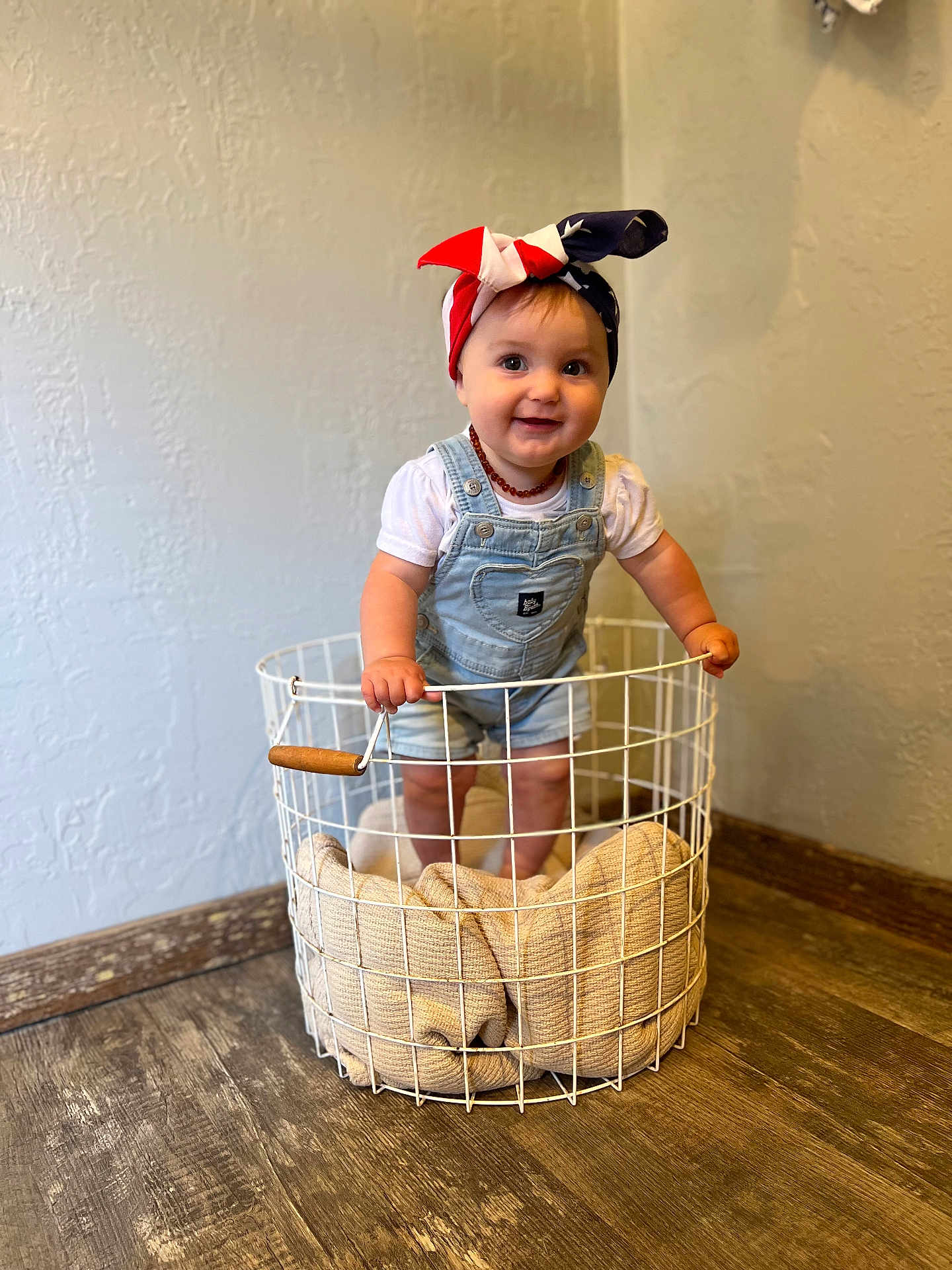 Lucy joined the competition — help win amazing prizes! baby, child, smiling, overalls, headband, basket, blanket, indoor, wooden_floor, wall, cute, happy, standing, portrait, clothing, person, infant, floor, headwear, expression