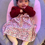 Melek is registered to the contest to win money with this photo: child, baby, toddler, portrait, sitting, baby_seat, smiling, face, red_bow, dark_hair, sweater, floral_dress, striped_tights, pink_cushion, indoor, wooden_floor, rug, furniture, finger_in_mouth, cozy